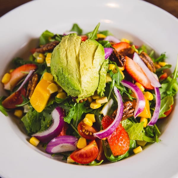 Colorful mixed green salad topped with avocado, corn, tomatoes, strawberries, red onion, and grilled vegetables.