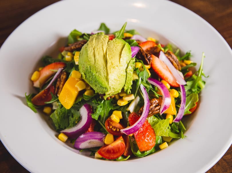 A vibrant salad with sliced avocado, strawberries, red onion, corn, and mixed greens in a white bowl.