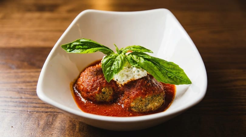 Two meatballs in red sauce topped with ricotta cheese and fresh basil in a white bowl.