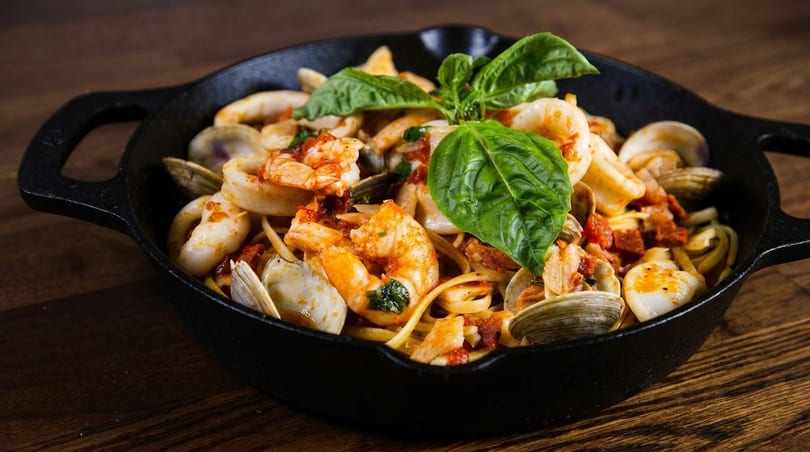 Seafood linguine with shrimp, clams, squid, and basil in a black cast iron pan.