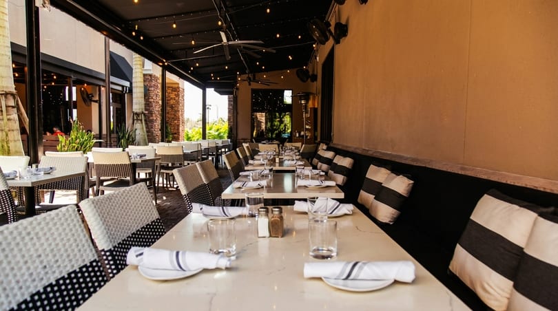 Outdoor dining patio with set tables, black and white striped cushions, and string lights under a dark awning.