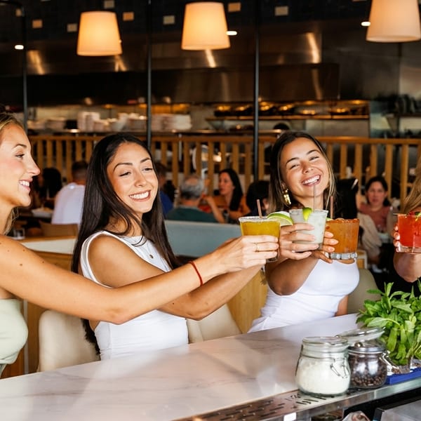 Four women smiling and toasting colorful cocktails at a lively restaurant bar.
