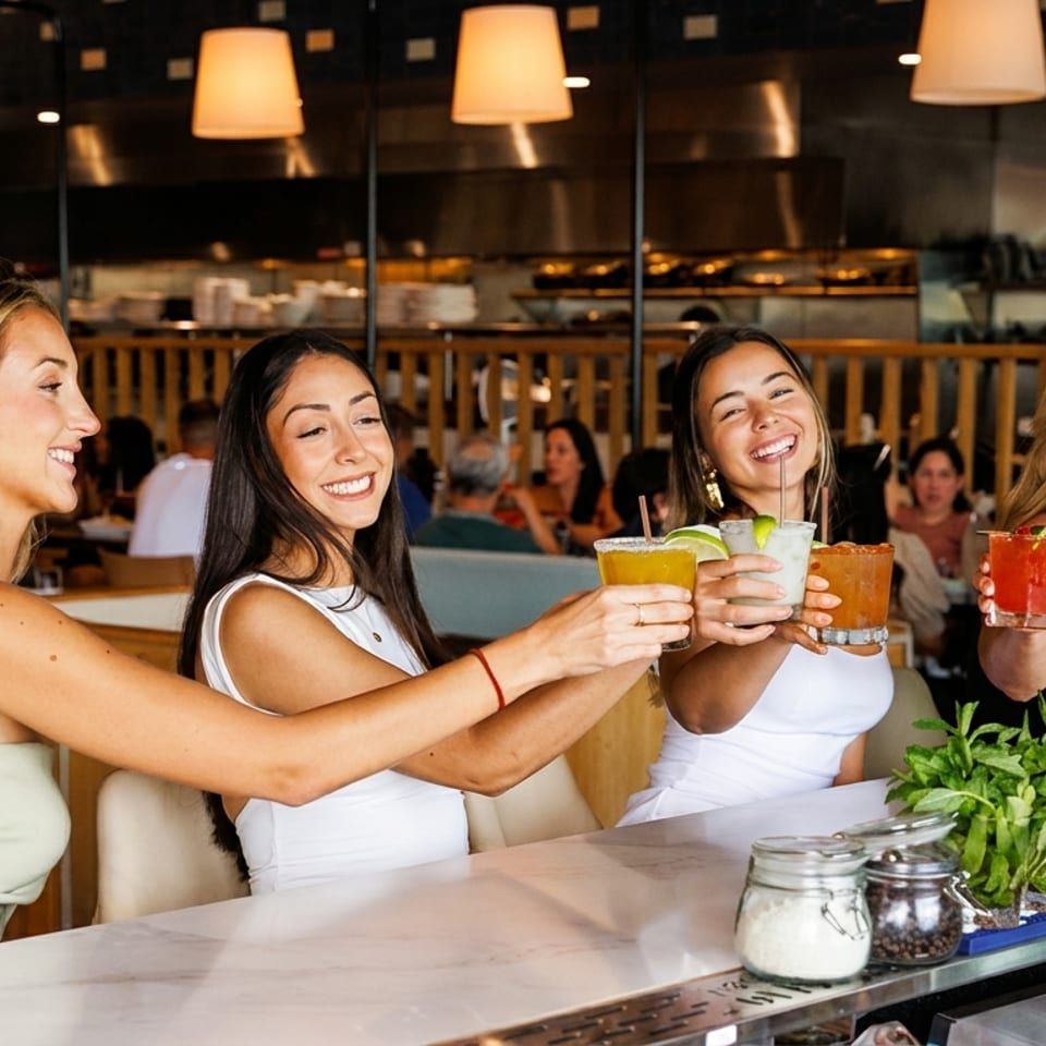 Four women smiling and toasting colorful cocktails at a lively restaurant bar.