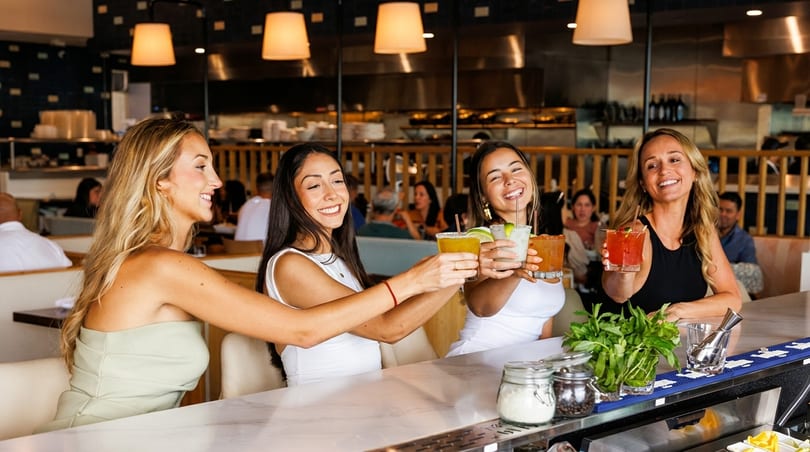 Four women smiling and toasting colorful cocktails at a lively restaurant bar.