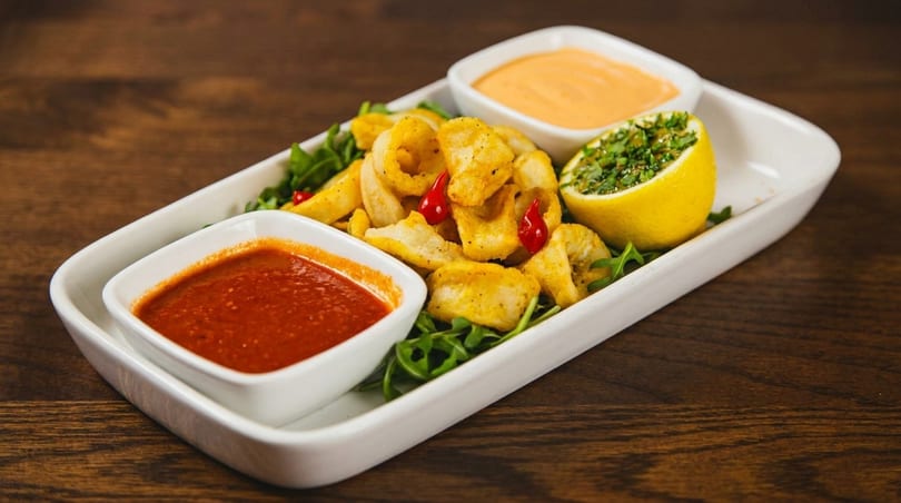 Fried calamari with two dipping sauces, red chili peppers, and a lemon half on a white platter.