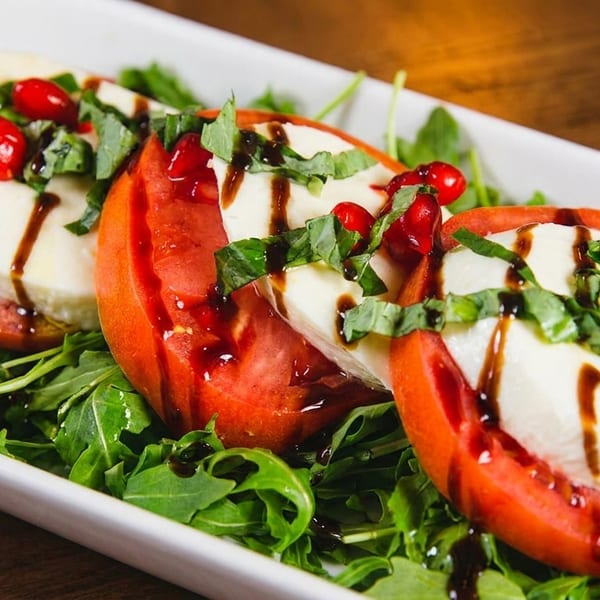 Three Caprese salad servings with tomatoes, mozzarella, basil, and balsamic glaze on a bed of arugula.