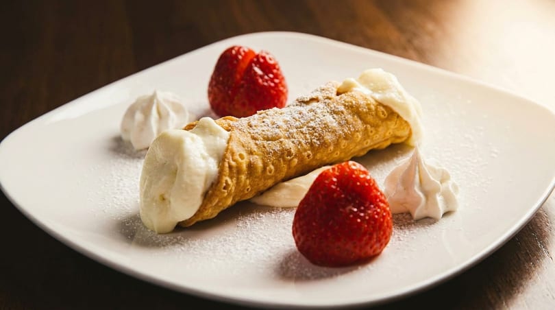 A cannoli dusted with powdered sugar, served with fresh strawberries and whipped cream on a white plate.