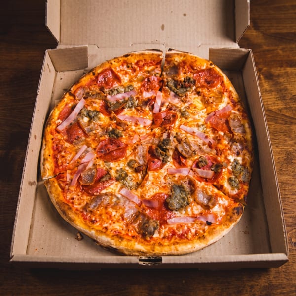 Loaded pepperoni, sausage, and ham pizza in an open cardboard box on a wooden table.