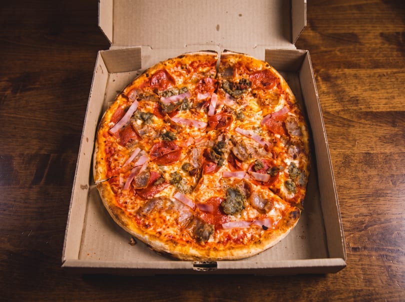 A large, sliced pizza topped with pepperoni, ham, and ground meat in an open cardboard box.