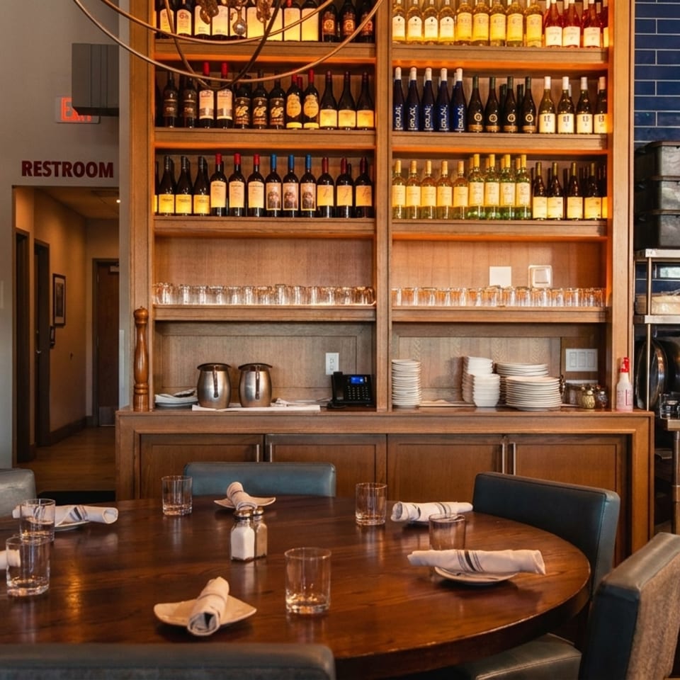 Restaurant interior with a round wooden table, wine shelves, and a kitchen area with blue tiled walls.