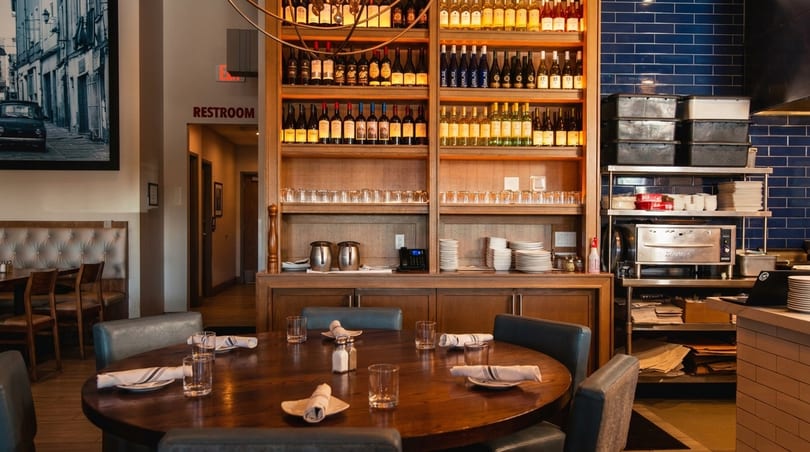 Restaurant interior with a round wooden table, wine shelves, and a kitchen area with blue tiled walls.