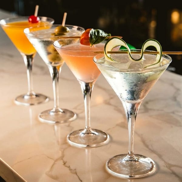 Four colorful cocktails in martini glasses with various garnishes lined up on a marble bar.
