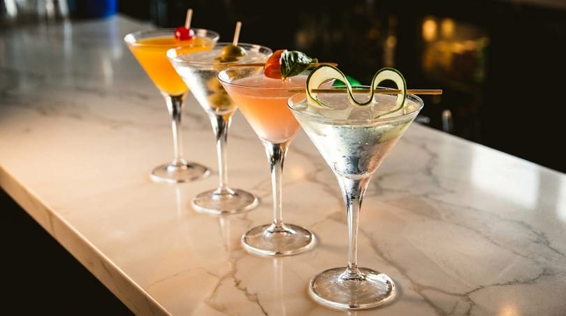Four colorful cocktails in martini glasses with various garnishes lined up on a marble bar.