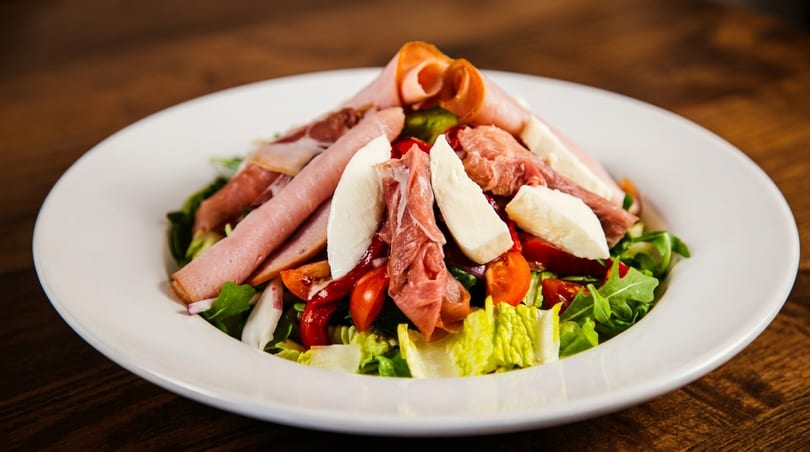 A fresh salad with mixed greens, cherry tomatoes, mozzarella, and various deli meats on a white plate.