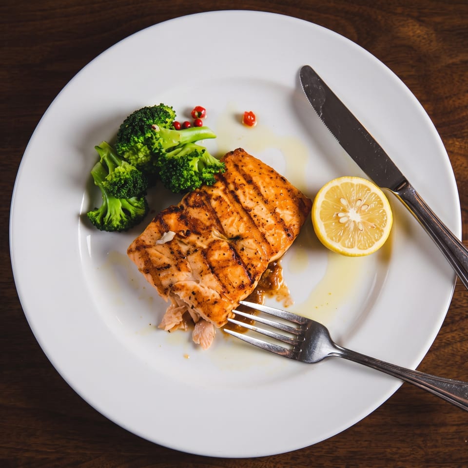 Grilled salmon fillet served with steamed broccoli and lemon on a white plate, accompanied by a fork and knife.