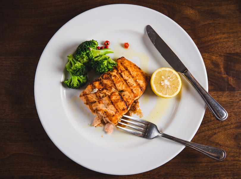 Grilled salmon with broccoli, a lemon slice, fork, and knife on a white plate.