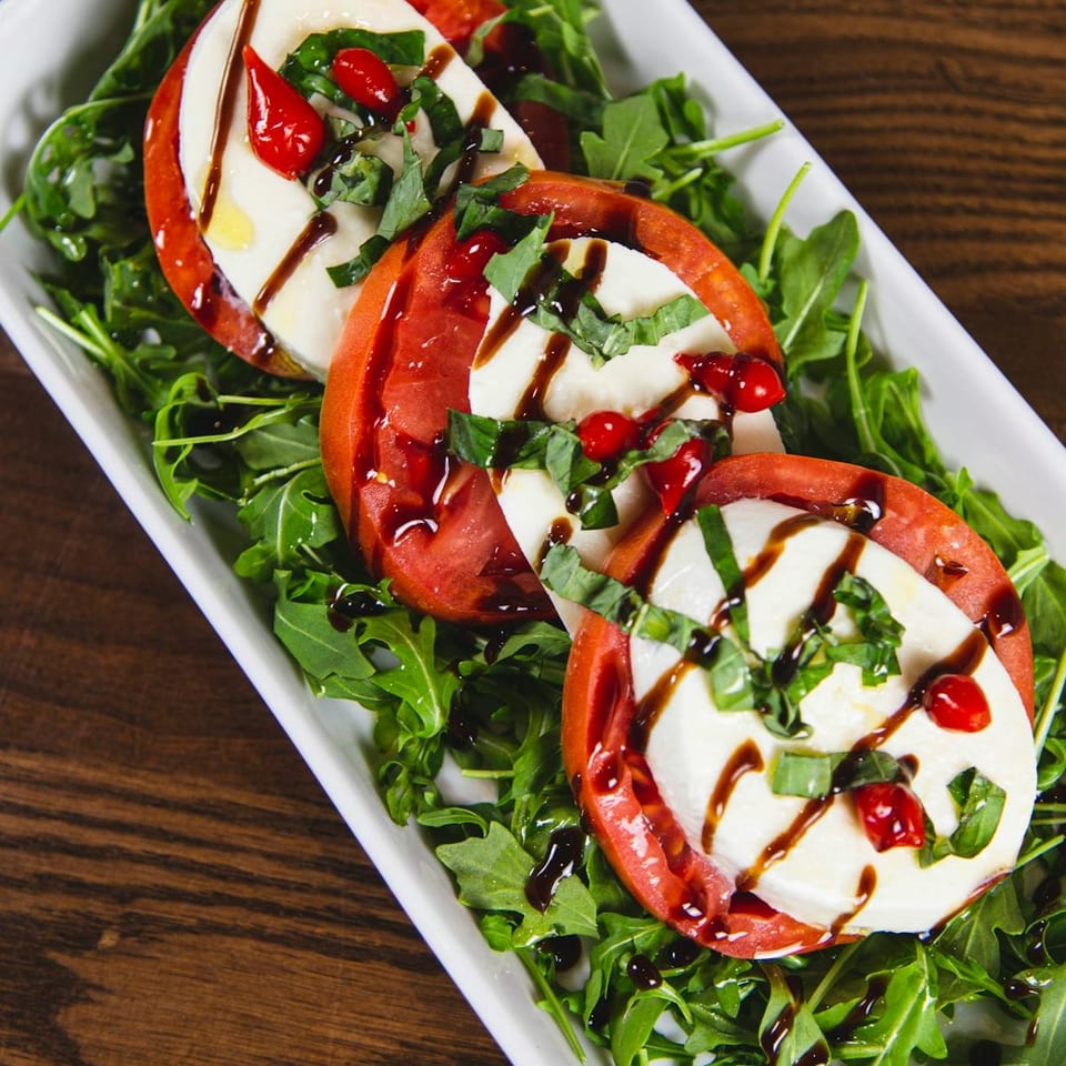 Caprese salad with sliced tomatoes, mozzarella, basil, red peppers, and balsamic glaze on arugula.
