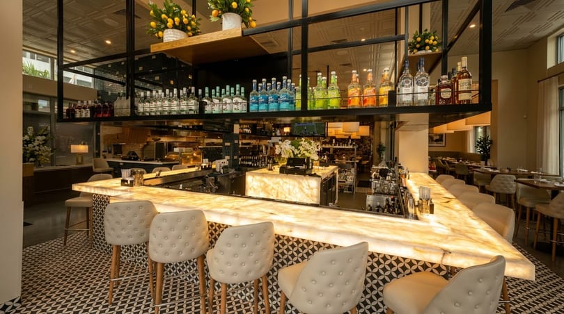 A modern restaurant bar with a glowing marble countertop, stylish bar stools, and liquor bottles on shelves.