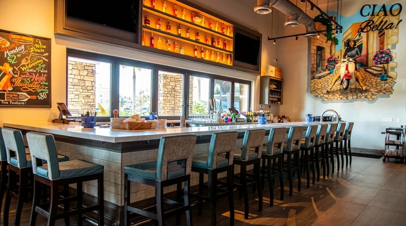 A long, well-lit bar with stools, a large window, and a "Ciao Bella!" mural.